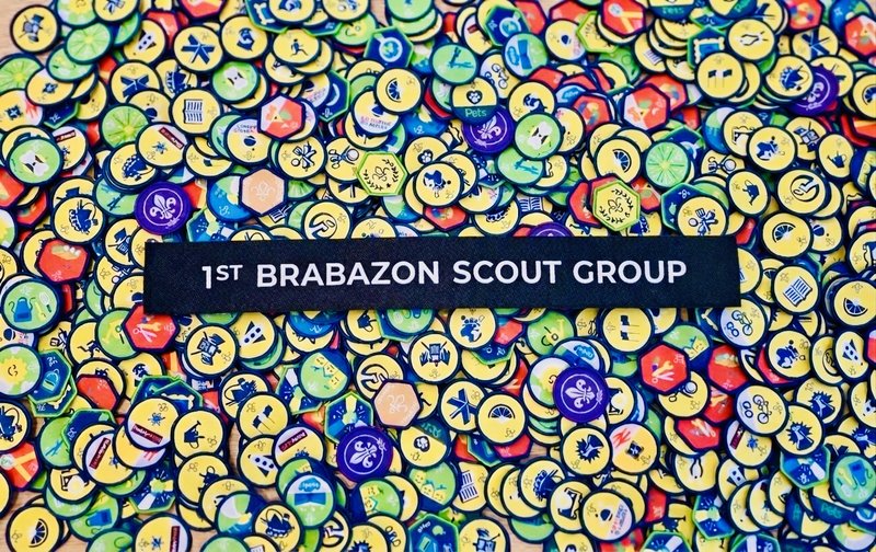 A woven 1st Brabazon Scout Group name tape rests on a colourful pile of Scout activity badges in yellow, green, blue and red.