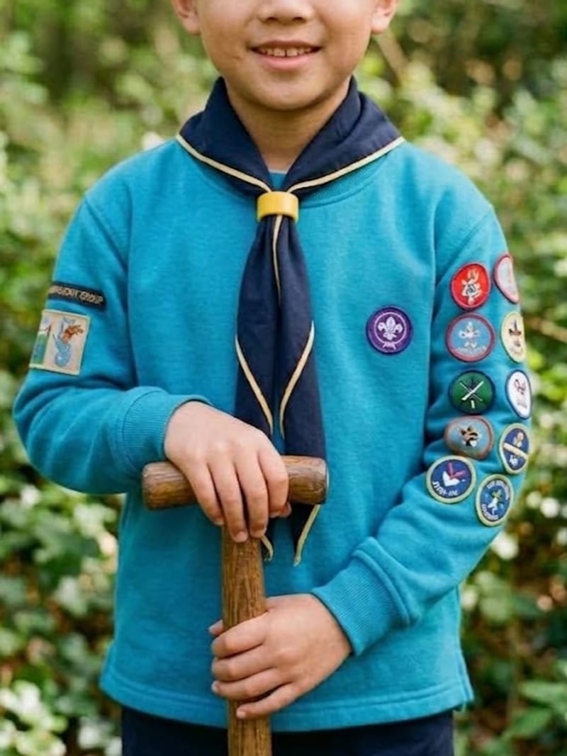 A Beaver Scout wearing the blue jumper and navy-and-gold group neckerchief, with badges sewn along the sleeve, standing outdoors.