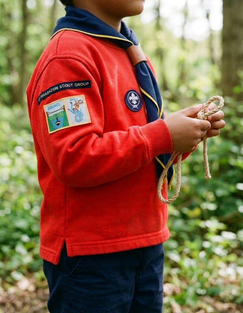 A Squirrel Scout in a red jumper and navy neckerchief coils a length of rope, standing among green woodland.